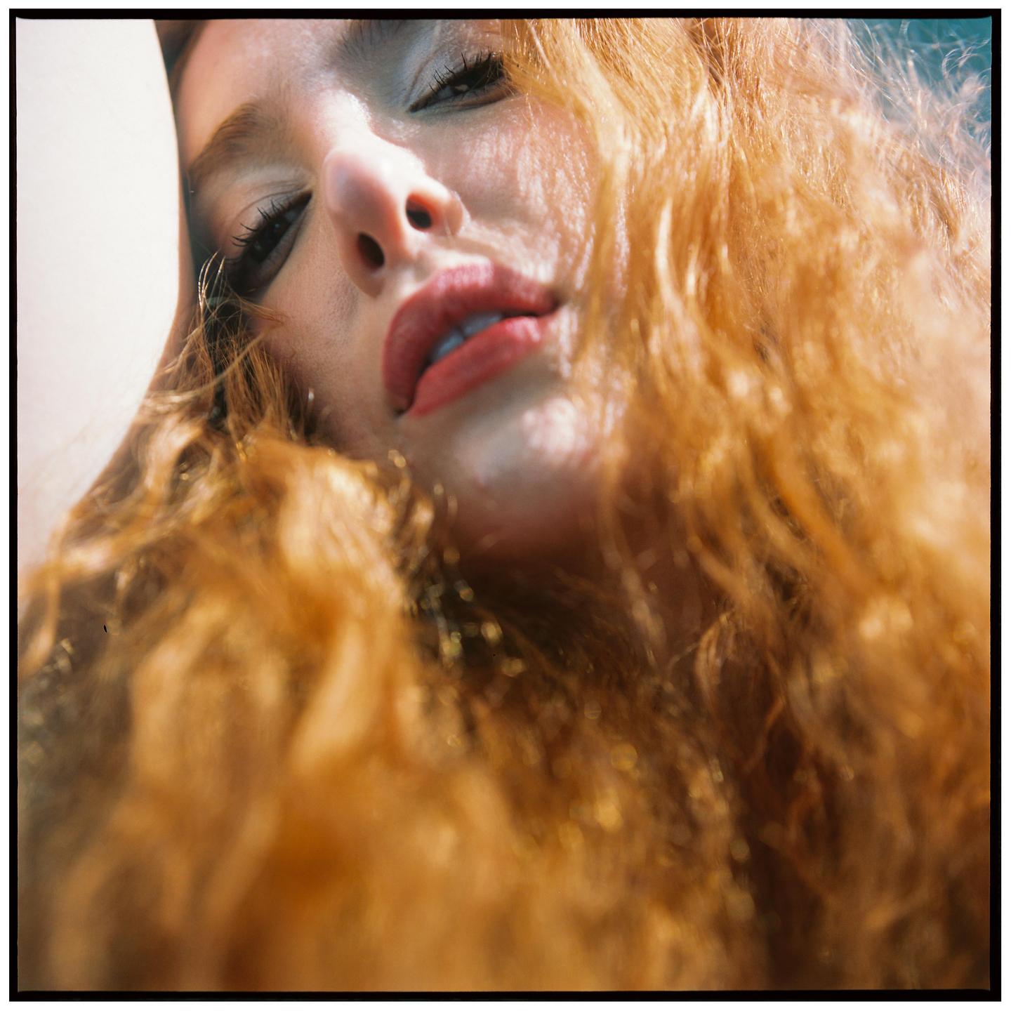 A woman with long, curly red hair and red lipstick looks up at the viewer.