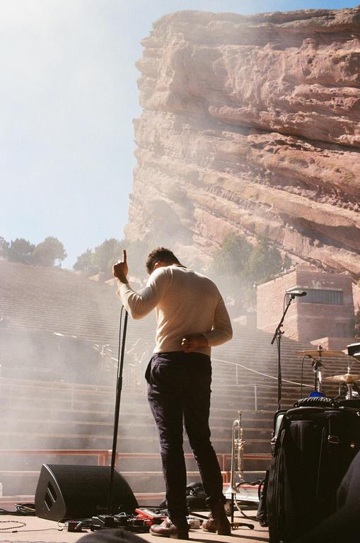 A man stands on a stage with a microphone, facing away from the viewer, with a large rock formation in the background.