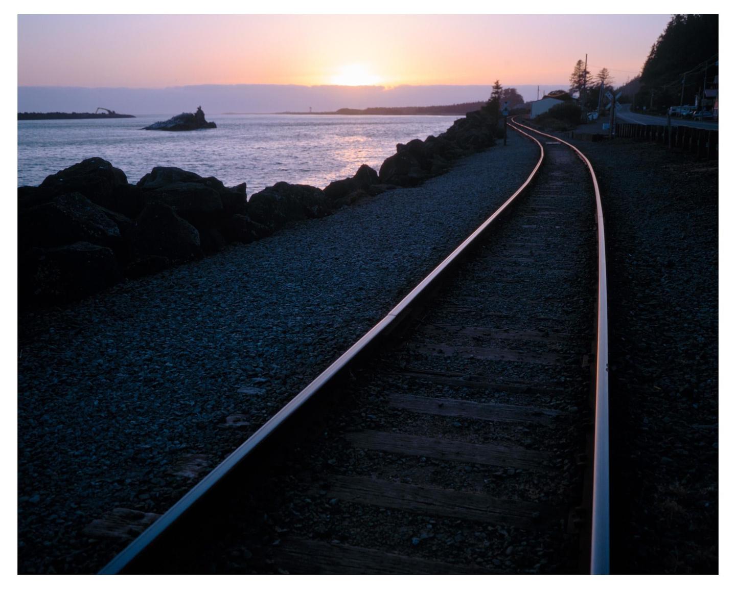 A railroad track curves along a rocky coast at sunset.