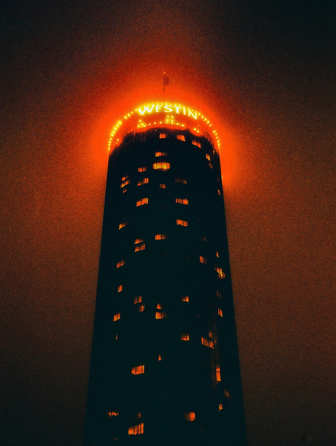 A tall, dark building with the word "WESTIN" illuminated in orange at its top stands against a hazy, dark sky.