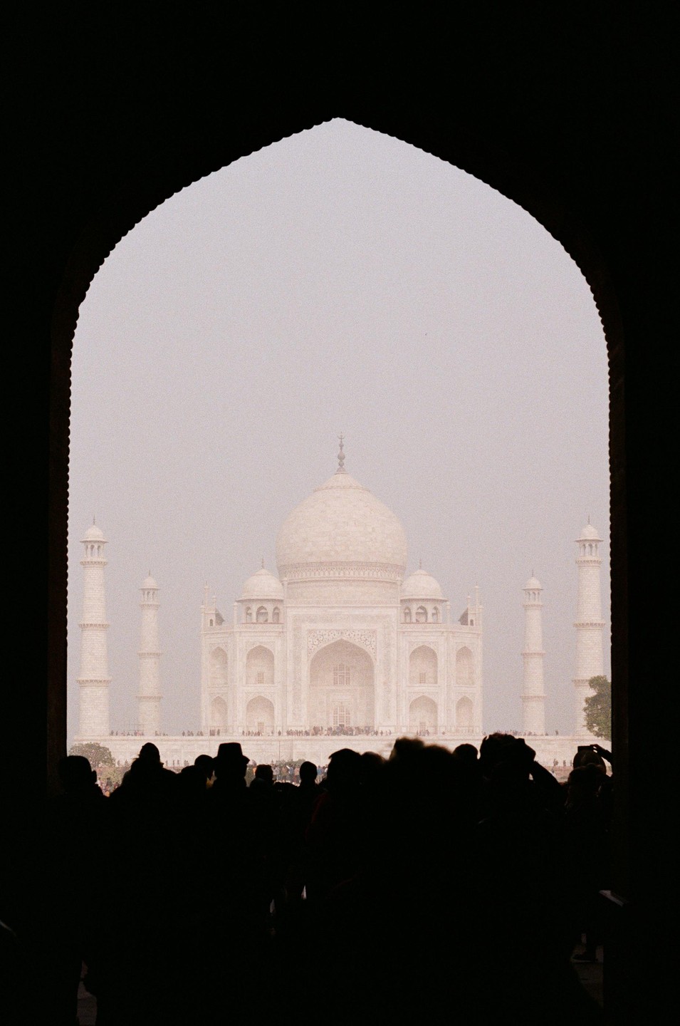 The Taj Mahal is framed by an archway with a crowd of people in silhouette in the foreground.