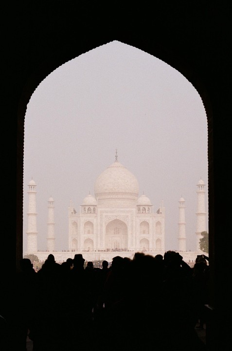 The Taj Mahal is framed by an archway with a crowd of people in silhouette in the foreground.