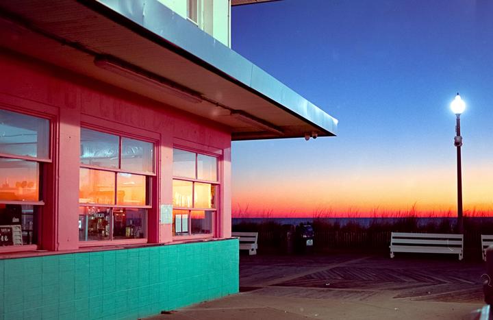 A pink building with a teal base stands next to a boardwalk with a bright orange and blue sunset over the ocean.