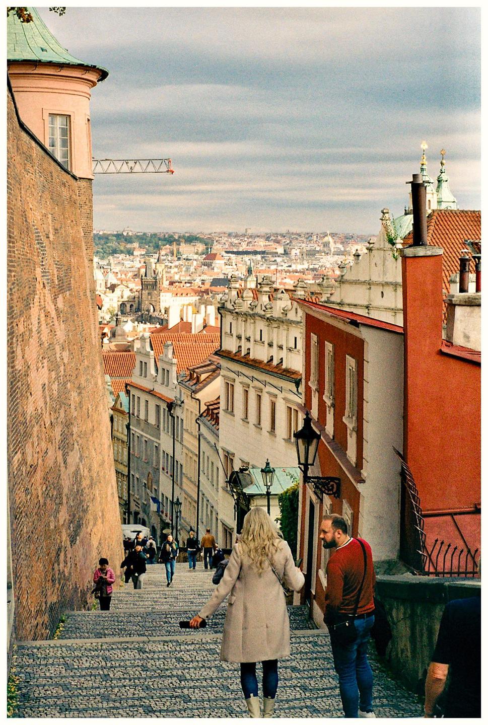A woman and a man stand on a cobblestone street looking down at the city of Prague.