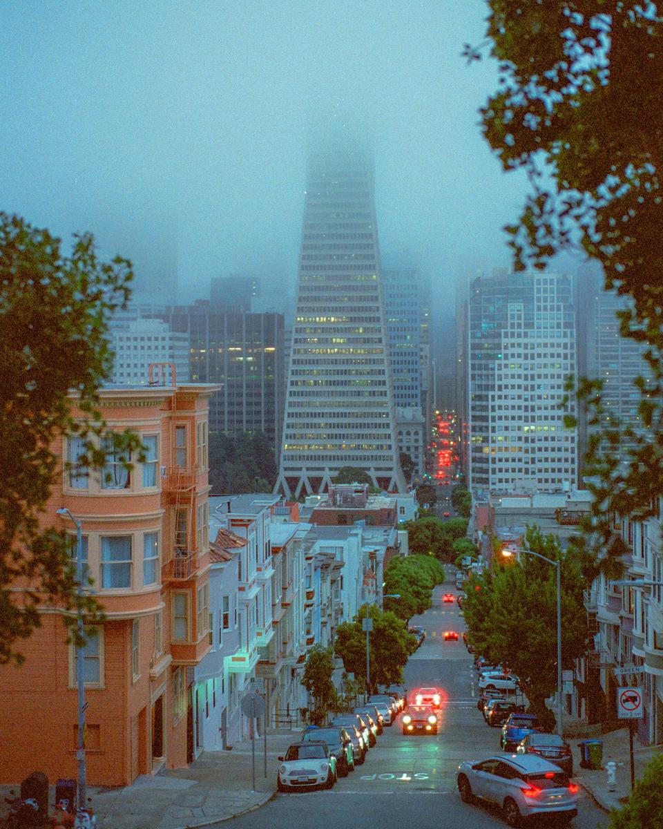 A foggy city street with parked cars and buildings on either side leads to a tall skyscraper partially obscured by fog.