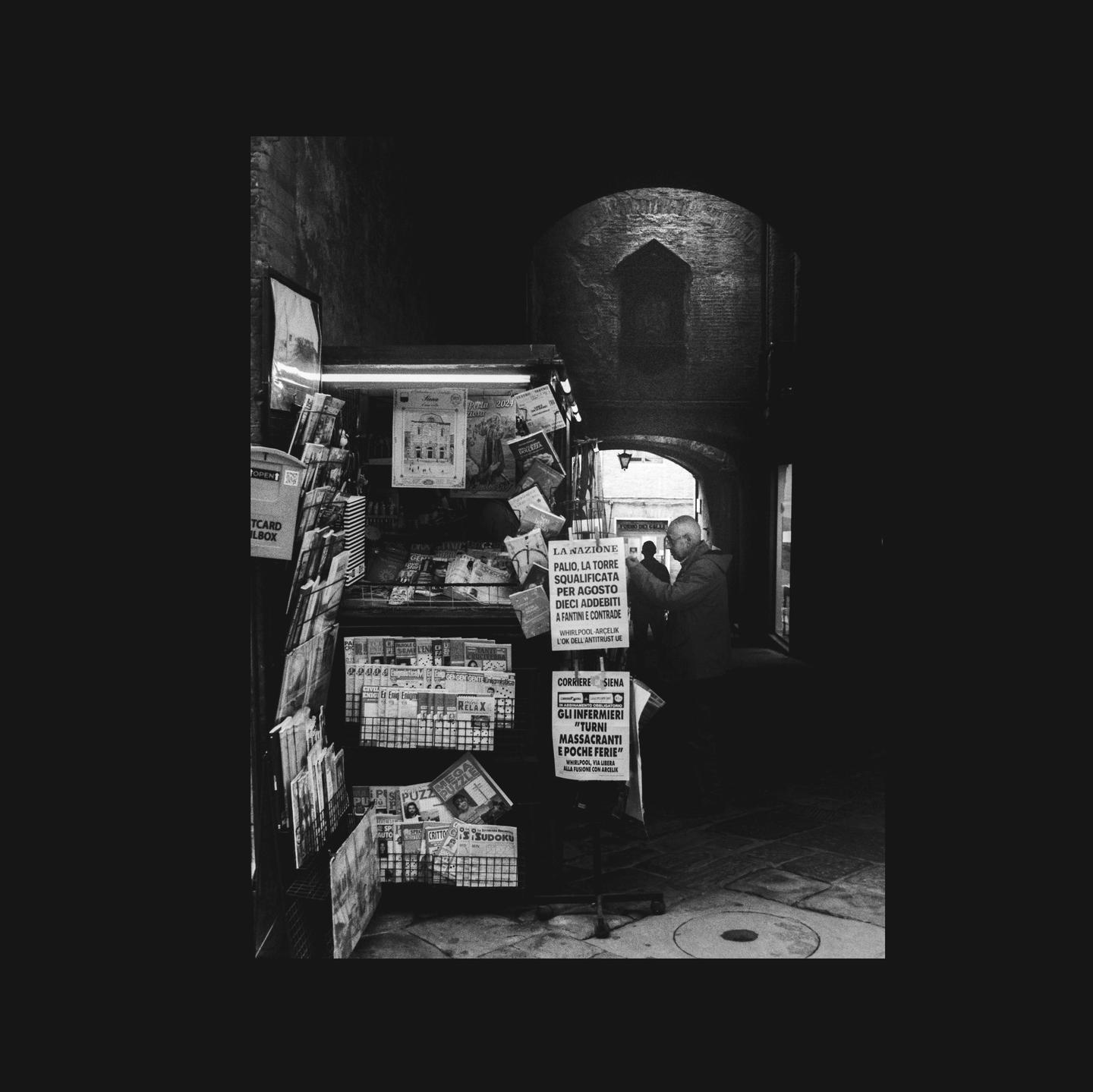 A man stands next to a newsstand in a dark alleyway in Siena, Italy.