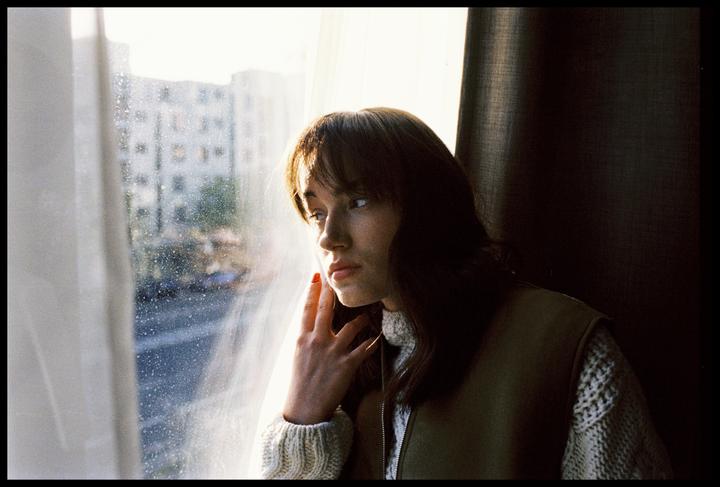 A woman with long hair and bangs looks out a window with her hand to her face.