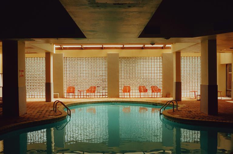 An indoor swimming pool with orange chairs and tables is surrounded by glass block walls.
