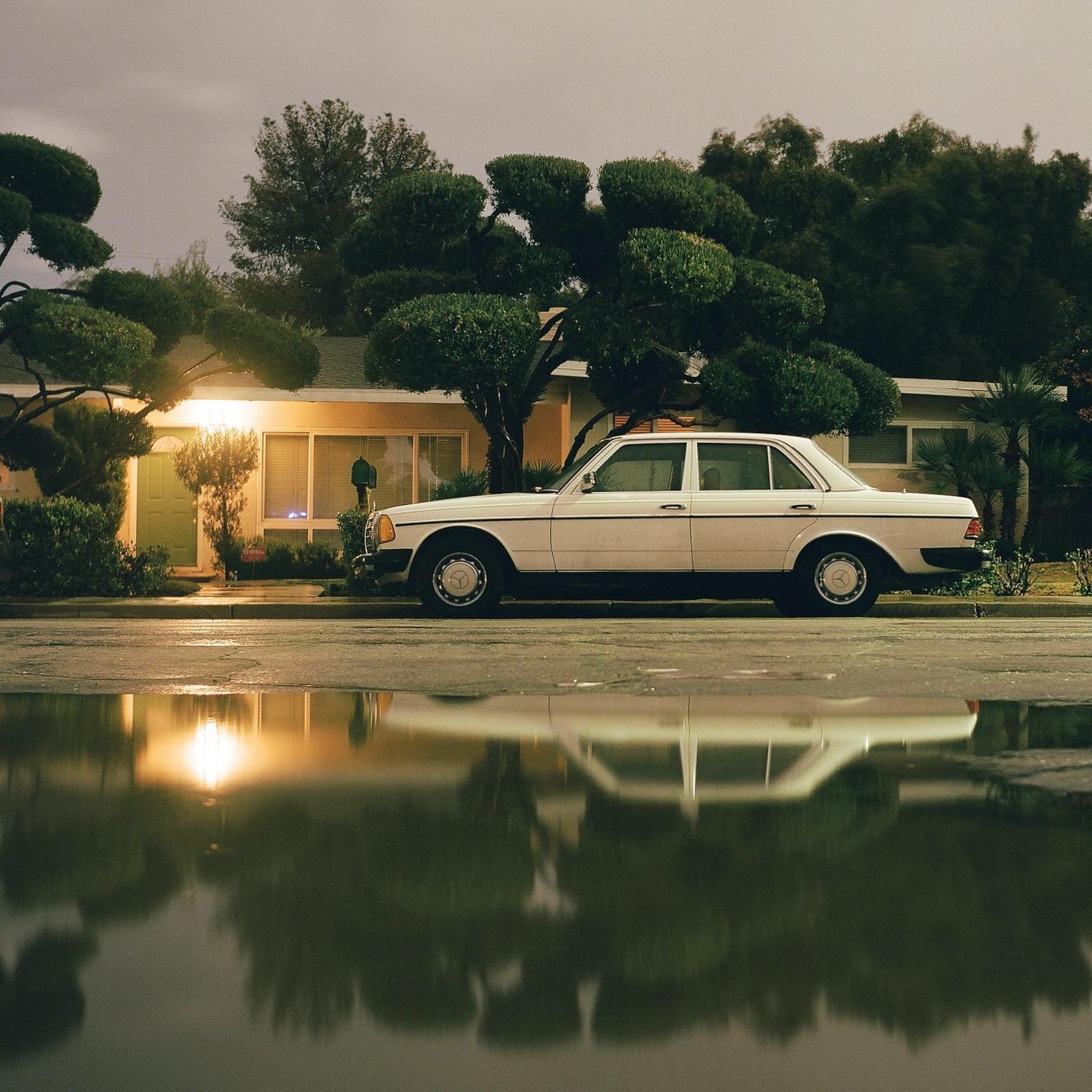 A white sedan is parked on a wet street in front of a house with manicured trees, reflecting in a large puddle.