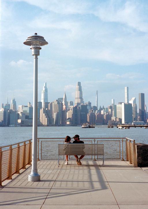 A couple sits on a bench by the water with a city skyline in the background.