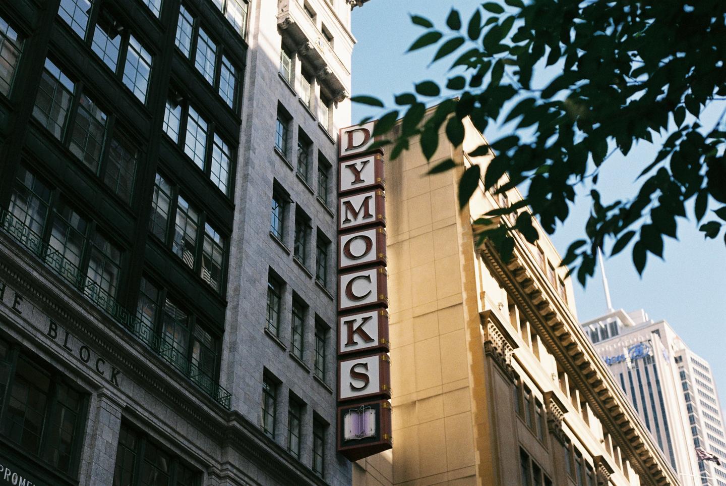 A tall, vertical sign for "Dymocks" is mounted on the side of a building, with other buildings and tree leaves visible.