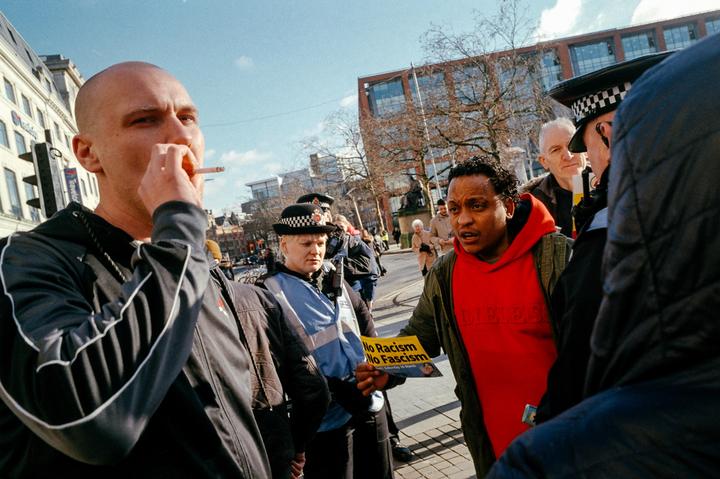 A man in a red hoodie holds a "No Racism No Fascism" sign while talking to a police officer on a city street.