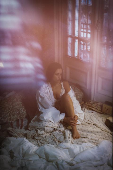 A woman sits on a bed in a sunlit room, looking down at her hands.