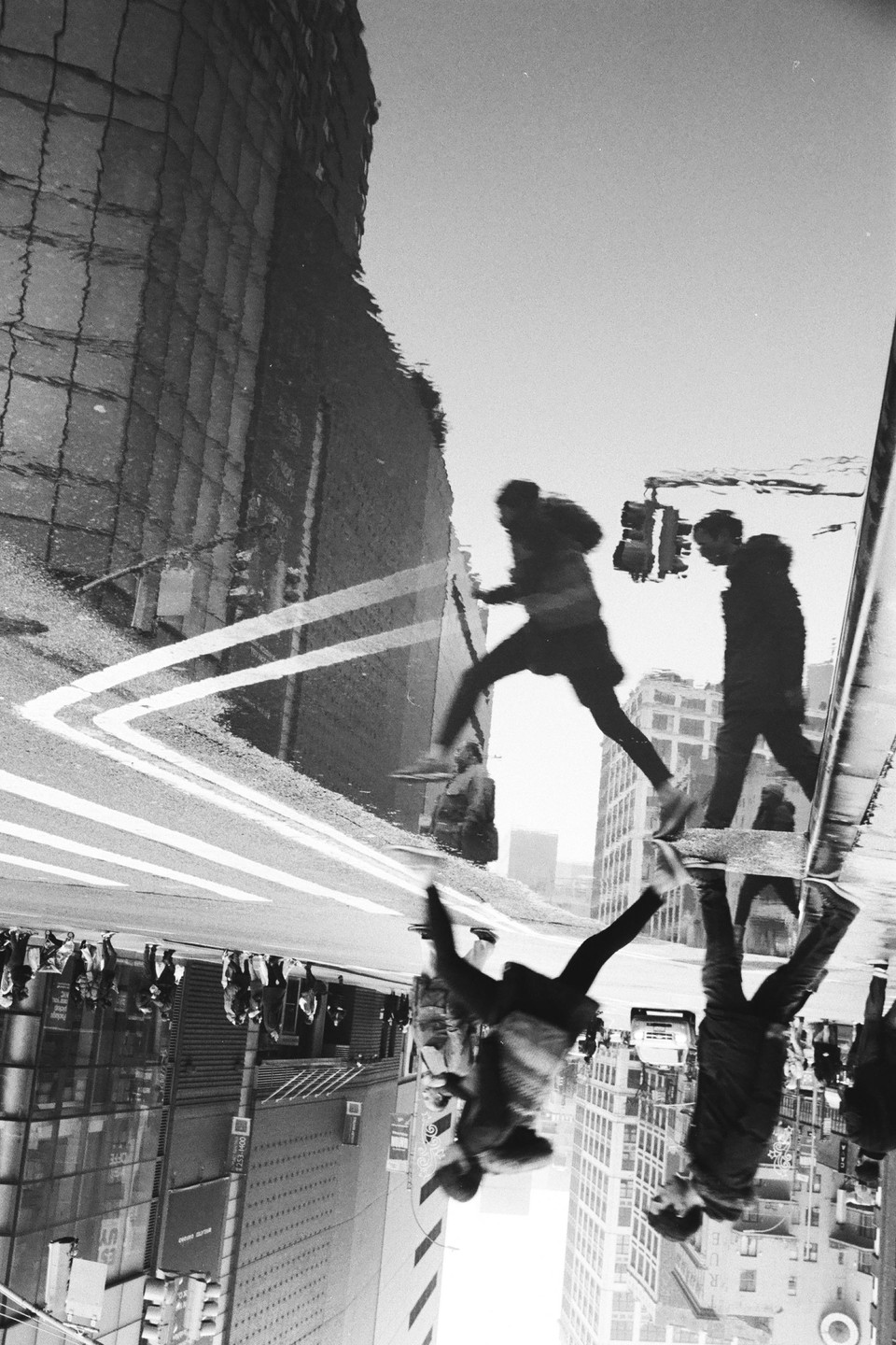 A man jumps over a puddle on a city street, with his reflection and the reflection of another man visible in the water.