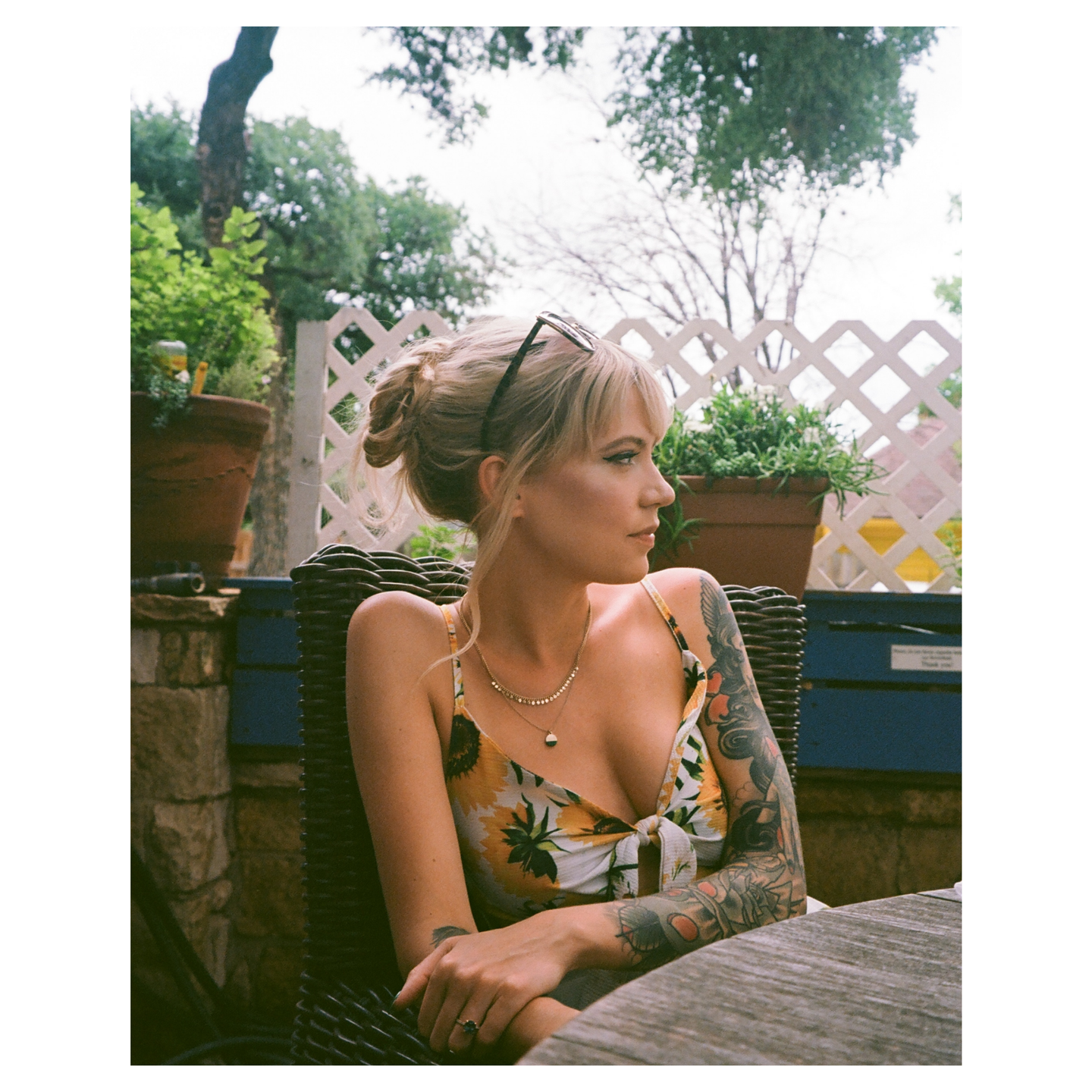 A woman with a sunflower-print top and tattooed arms sits at a wooden table outdoors, looking to her right.