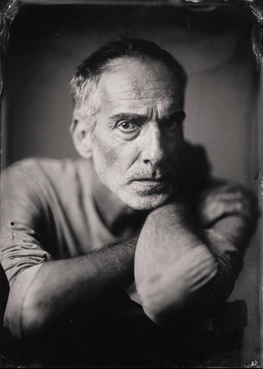A man with a beard and short hair leans forward with his arms crossed, looking directly at the viewer.