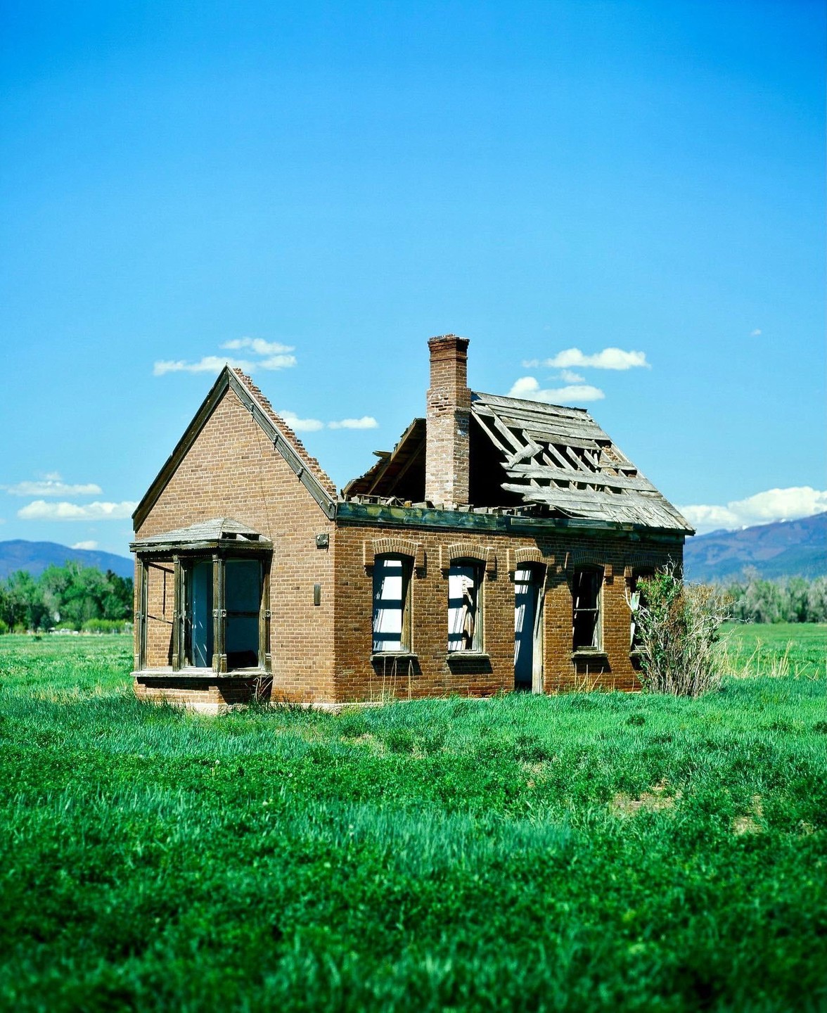 A dilapidated brick house with a collapsed roof stands in a vibrant green field under a clear blue sky.