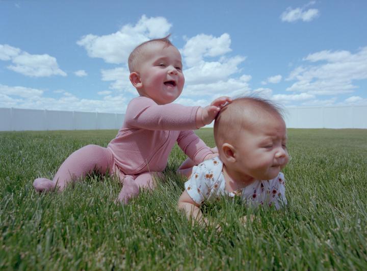 Two babies are playing in the grass under a blue sky with white clouds.