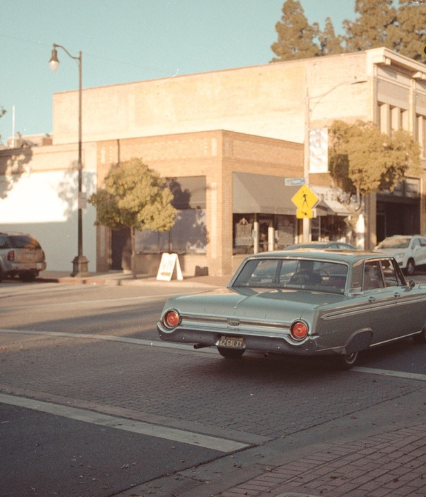 A vintage car is driving on a street in a town with brick buildings and trees.