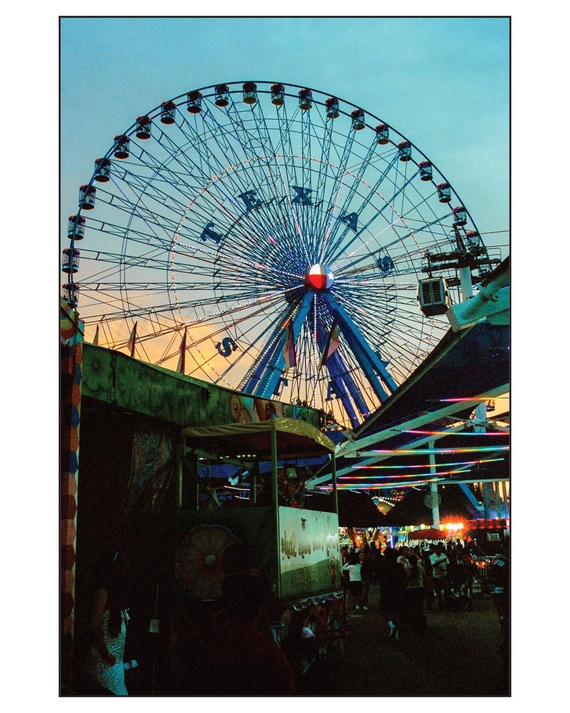 A large Ferris wheel with the word "TEXAS" spelled out in lights dominates the sky above a bustling fairground at dusk.
