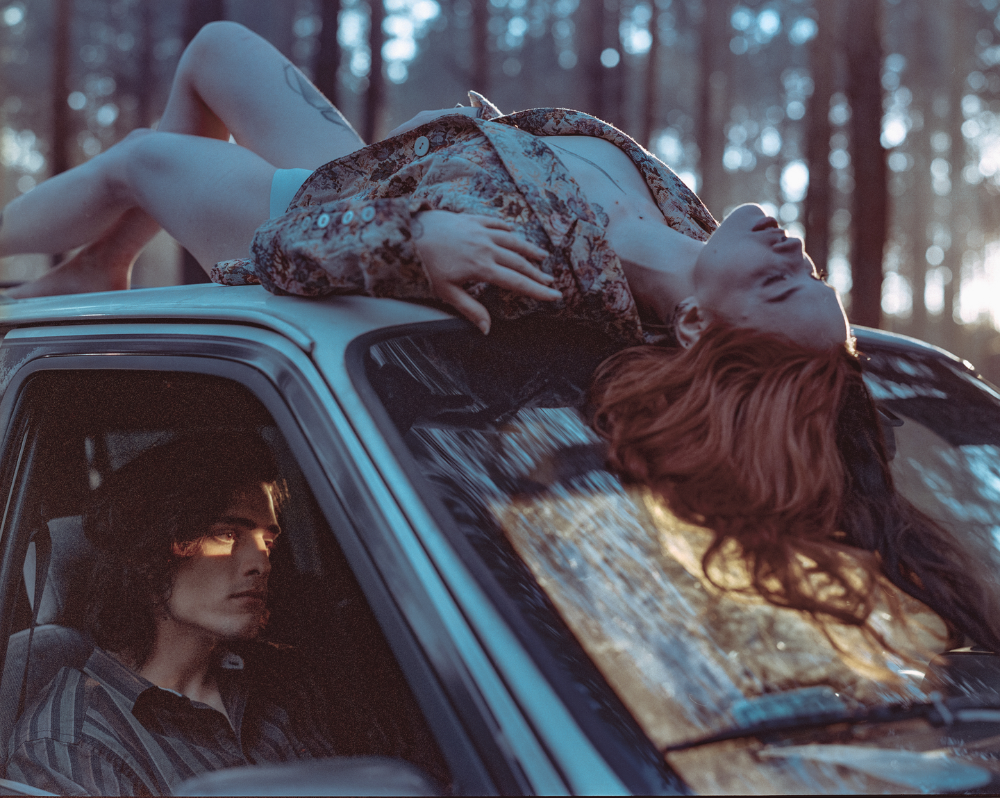 A woman lies on the roof of a car while a man sits in the driver's seat, with a forest in the background.