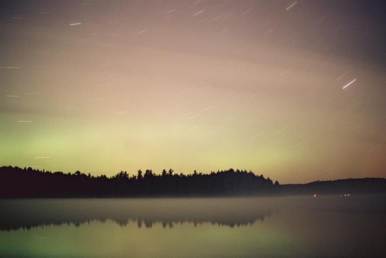 Star trails are visible above a forest reflecting in a misty lake at night.