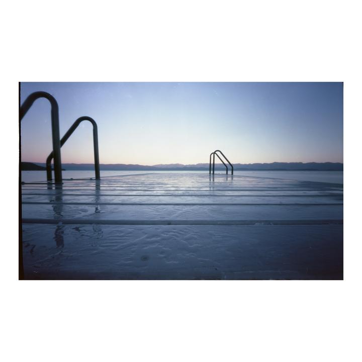 A wooden dock with two sets of metal steps leading into a body of water is shown under a clear sky with distant mountains.