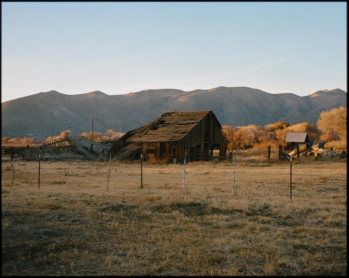 A dilapidated wooden barn stands in a dry field with mountains in the background.