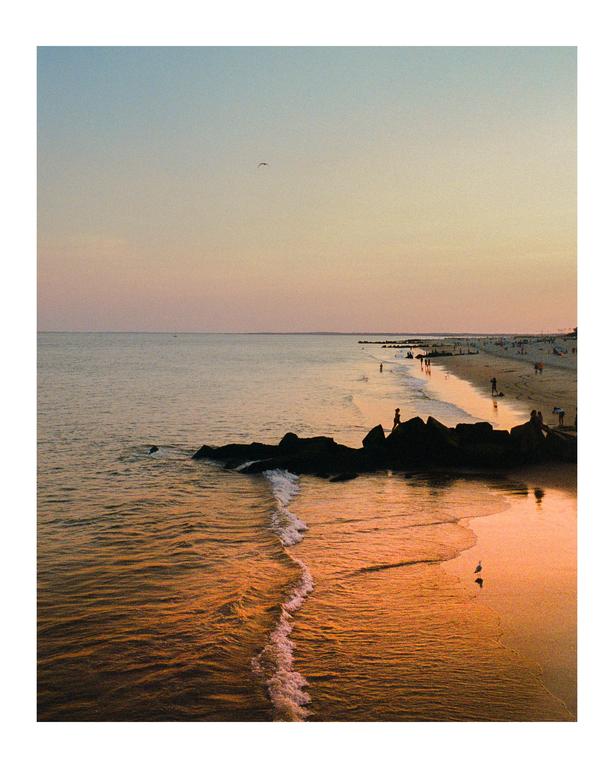 A beach is visible at sunset with people walking along the shore and rocks in the water.