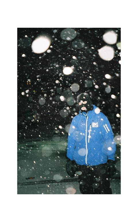 A man wearing a blue jacket and a black balaclava stands in falling snow at night.