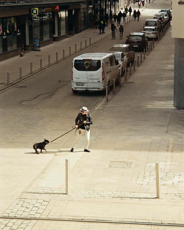 A person in a hat and sunglasses walks a dog on a long leash down a city street with parked cars and pedestrians.