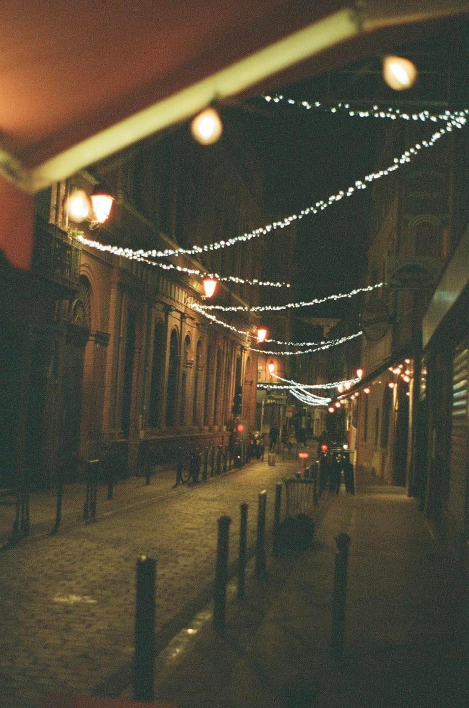 A narrow street at night is illuminated by strings of small lights strung across the buildings and a few streetlights.