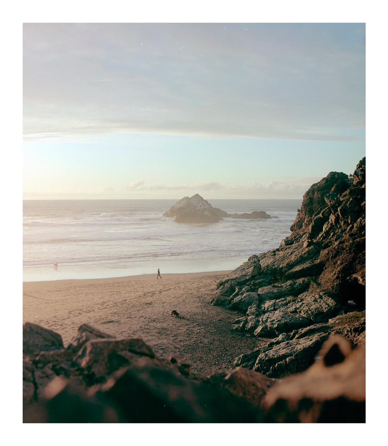 A person walks on a sandy beach next to a rocky cliff and ocean with a small island.