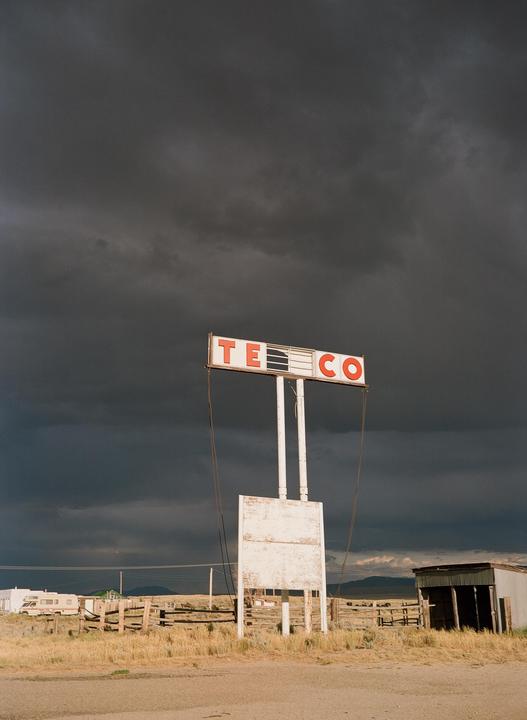 A weathered "TE CO" sign stands against a dramatic, dark cloudy sky in a rural landscape with a trailer and shed in the background.