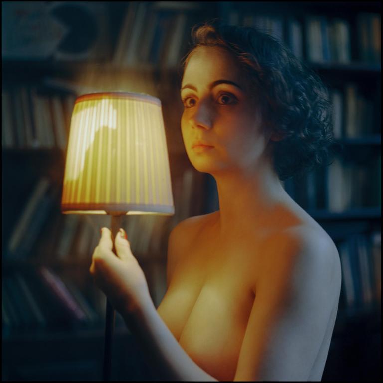 A woman holds a small lamp in front of a bookshelf.