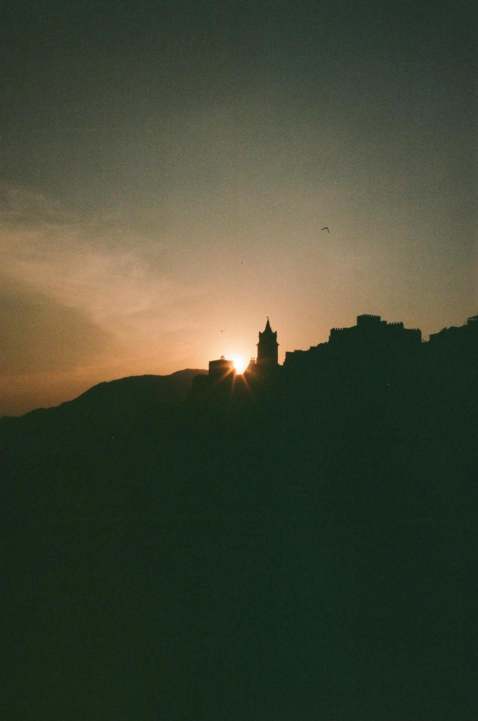 The sun sets behind a silhouetted town on a hill.