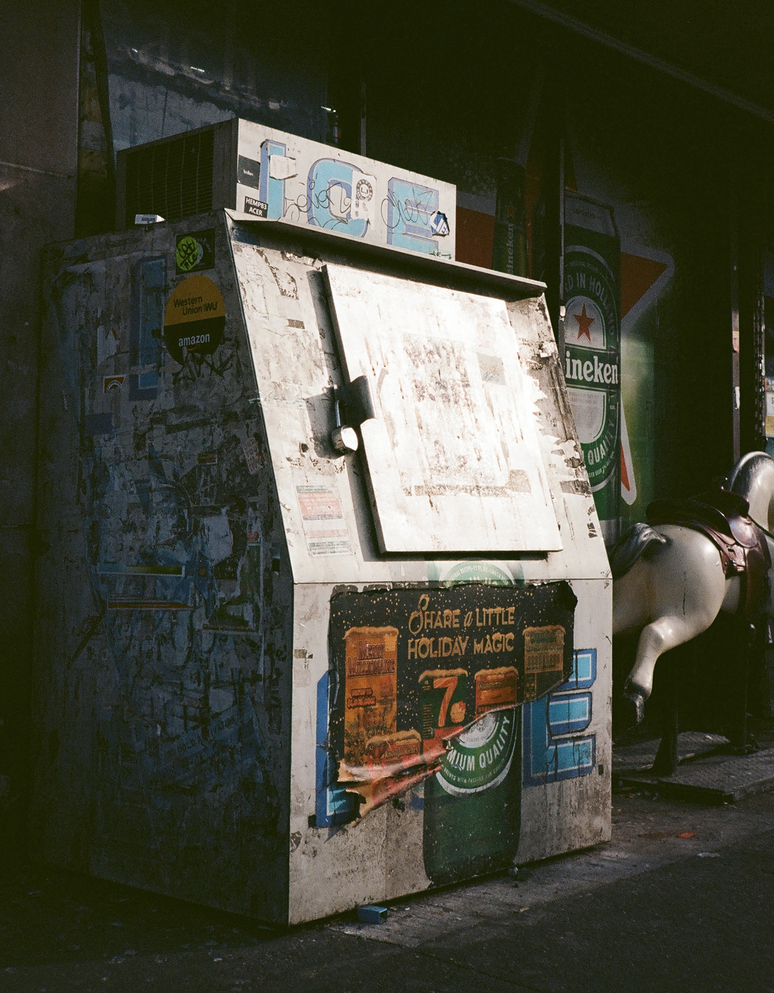 A weathered newspaper vending machine covered in stickers and advertisements stands next to a coin-operated horse ride.