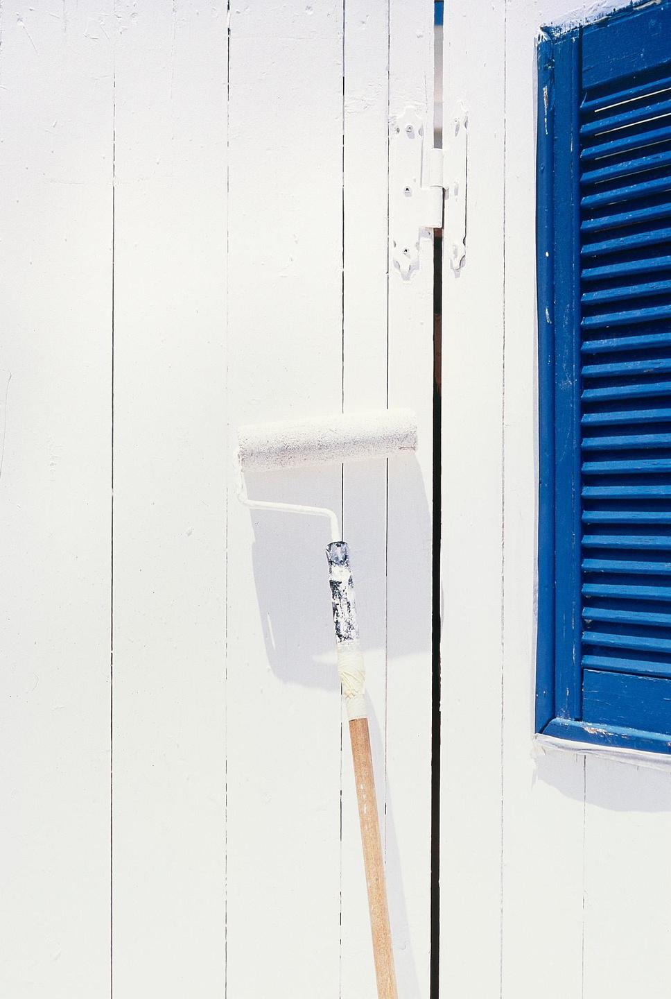 A paint roller with a long handle leans against a white wooden wall next to a blue shutter.