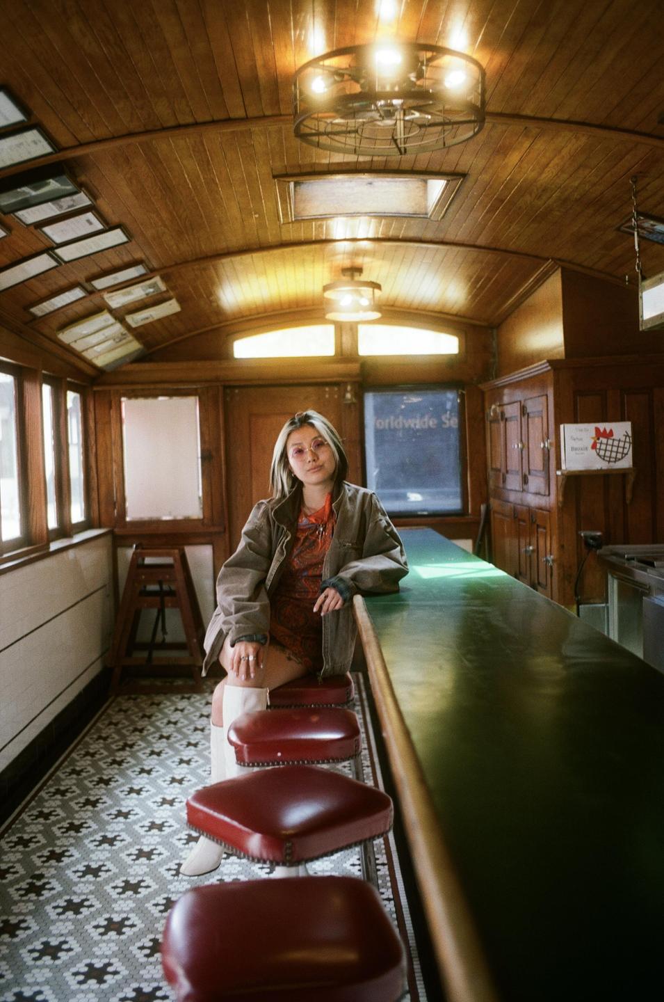 A woman sits on a stool at a long counter in a diner with wood paneling and patterned floor tiles.
