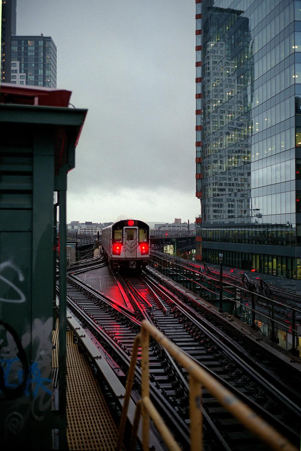 A subway train with red lights on its rear travels on elevated tracks next to a modern glass building under a cloudy sky.