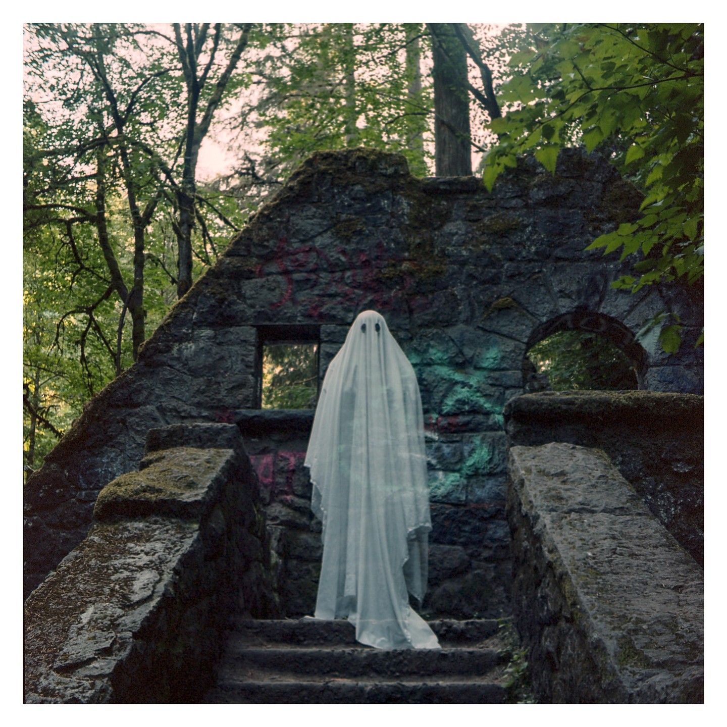 A person in a white sheet ghost costume stands on stone steps in front of a ruined stone building in a forest.