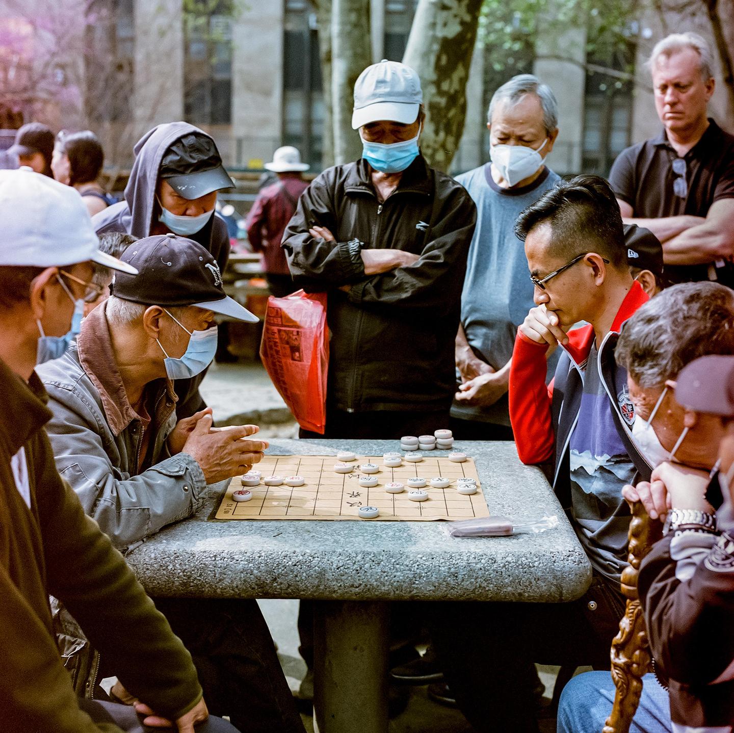 A group of men wearing face masks are gathered around a stone table playing a board game outdoors.