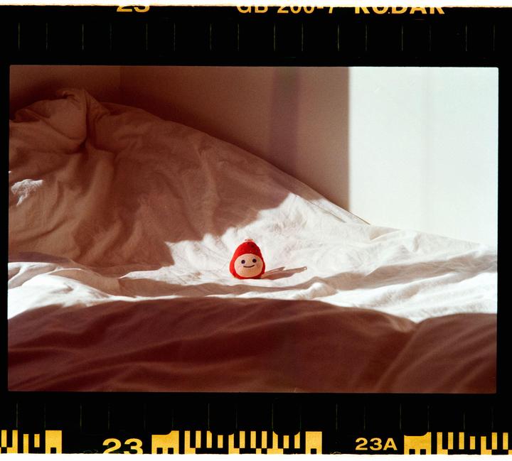 A small red doll with a smiling face sits on a white bedspread in a sunlit room.