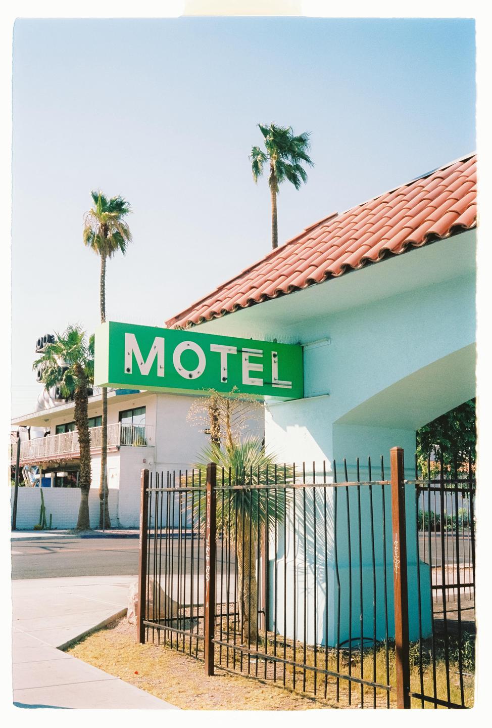 A green motel sign is visible above a black fence with palm trees and a building in the background.