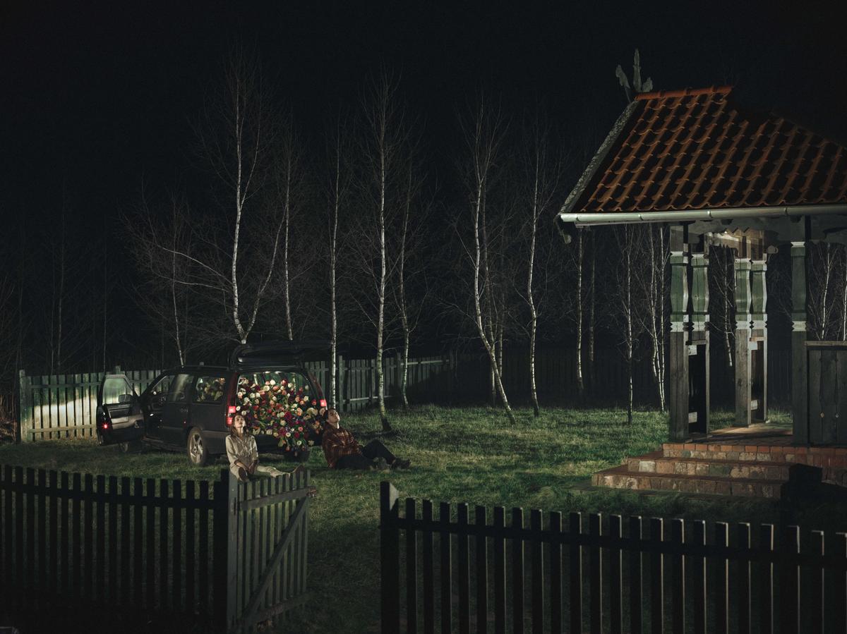 A couple sits on the grass next to a car filled with flowers at night, with a small building and trees in the background.