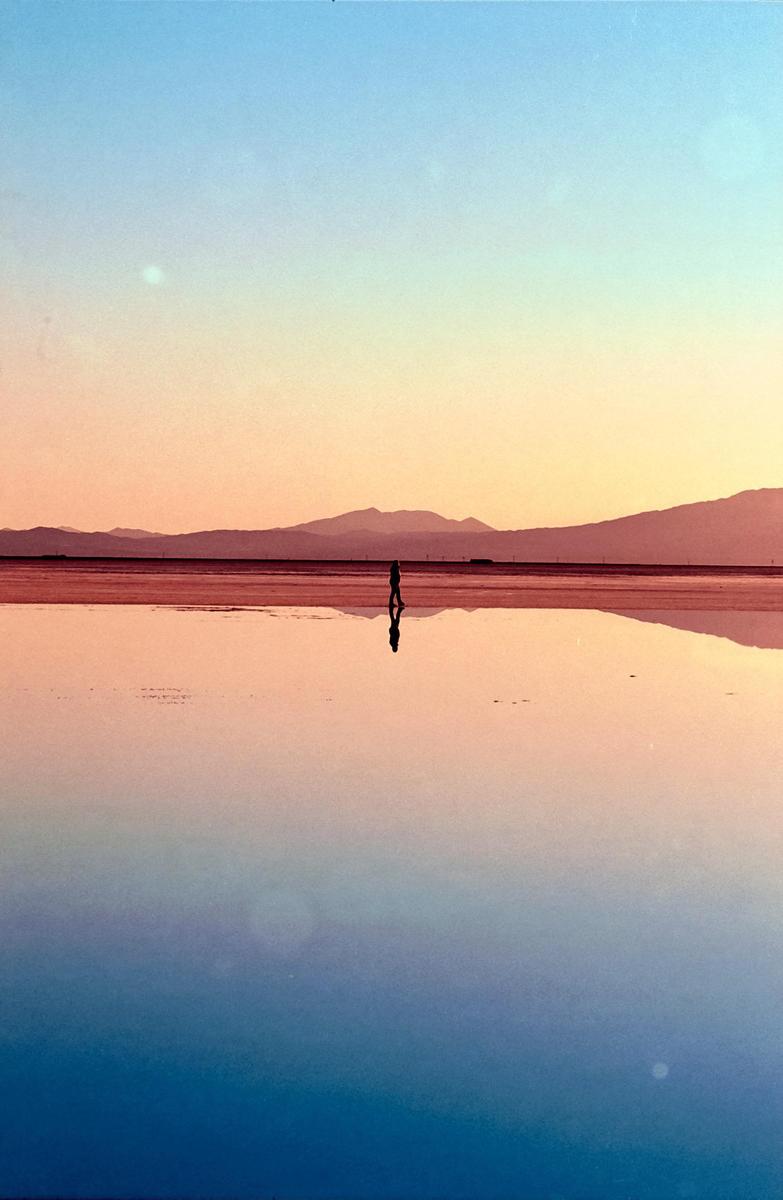 A person stands in a shallow body of water reflecting the colorful sky and distant mountains at sunset.