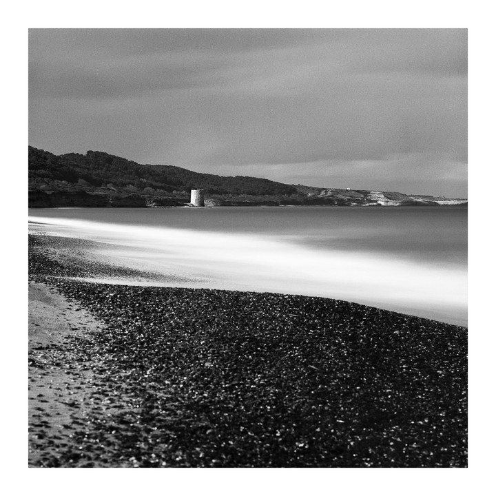 A long exposure photograph shows a beach with a tower in the distance and blurred water.