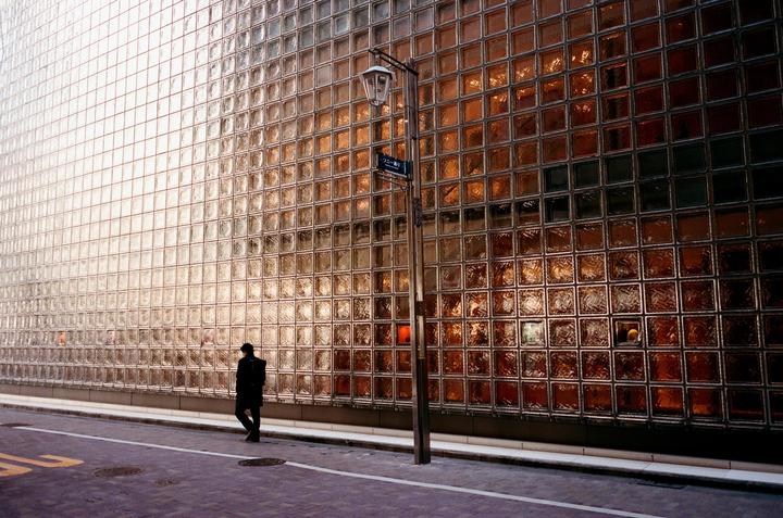 A person walks along a street next to a building with a glass block facade.