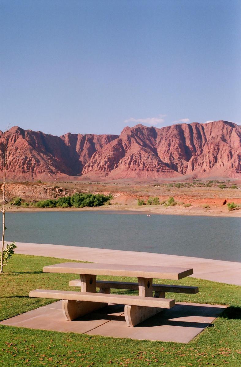 A picnic table sits on a grassy area next to a body of water with red mountains in the background.