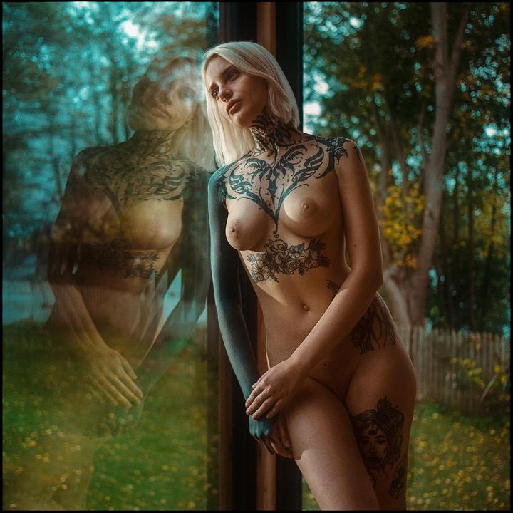 A nude woman with tattoos stands next to a window, with her reflection visible in the glass.